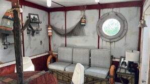 L’intérieur d’une cabine comprenant un canapé, une petite table avec une lampe, un hamac et une fenêtre ronde.