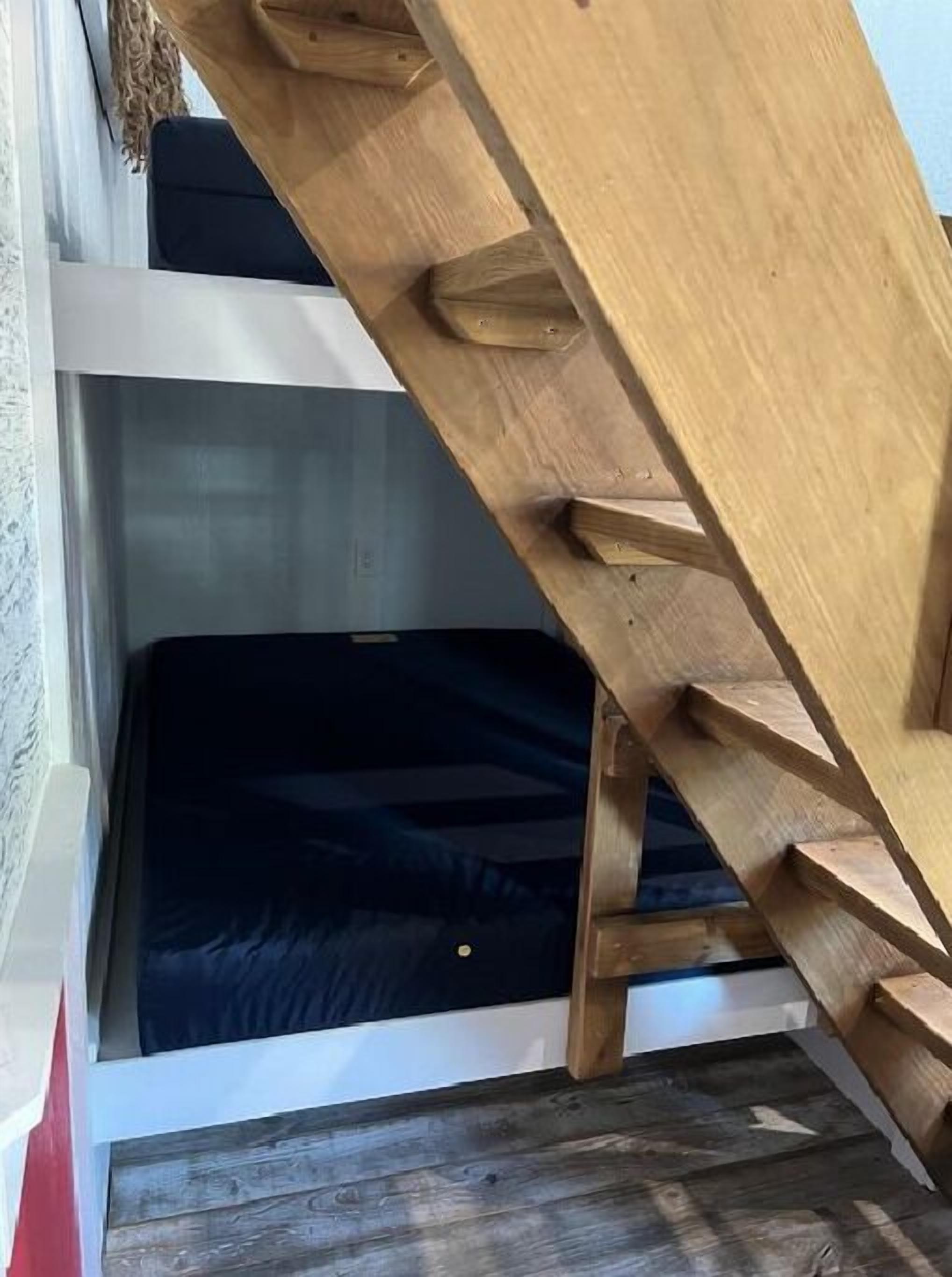 Un escalier en bois mène à un niveau inférieur où l’on aperçoit un matelas de couleur sombre.