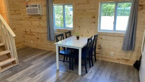 Una cabaña de madera con una mesa y sillas para comer, dos ventanas con cortinas, una escalera y un equipo de aire acondicionado.