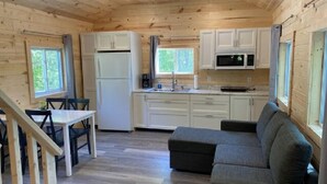 Cocina compacta con gabinetes blancos, un refrigerador y un microondas. Hay un sofá gris y una mesa de comedor con sillas.