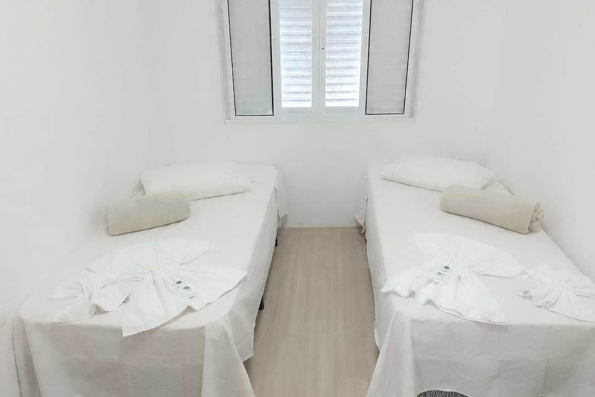 A small, clean room with two single beds, a window with blinds, and a plain white wall.