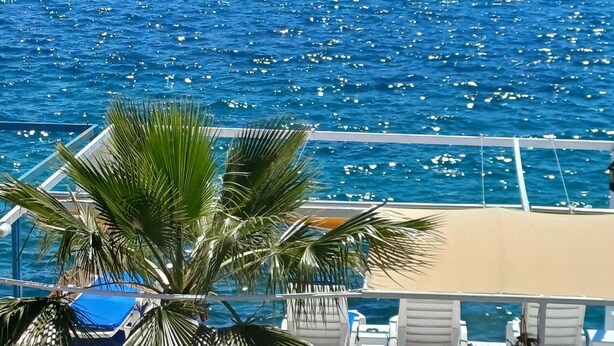 Eine Terrasse mit weißen Liegestühlen und einer Palme, mit Blick auf ein blaues Meer mit Booten.