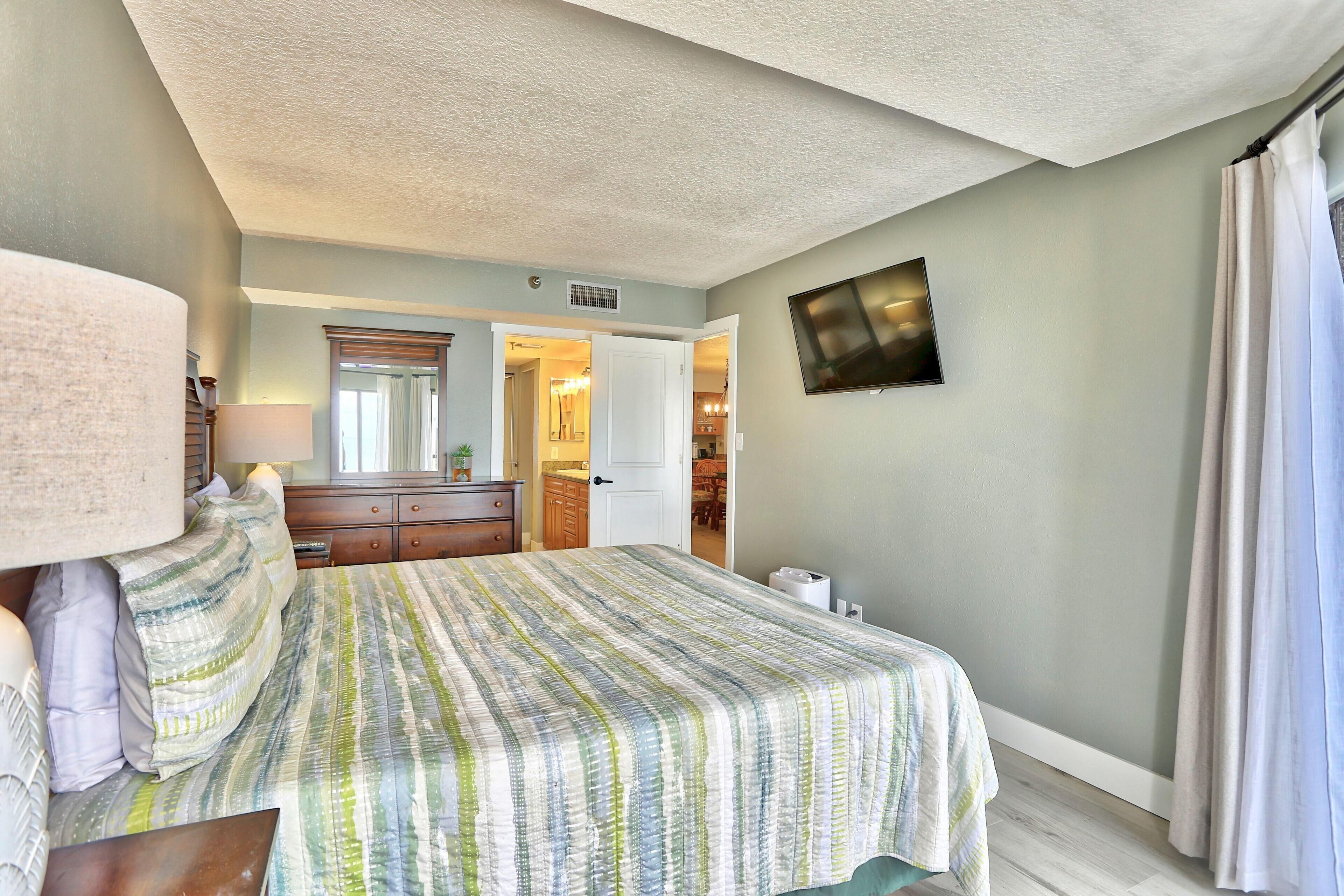 A bedroom with a bed, a TV mounted on the wall, a door leading to a bathroom, and a small table with a lamp.