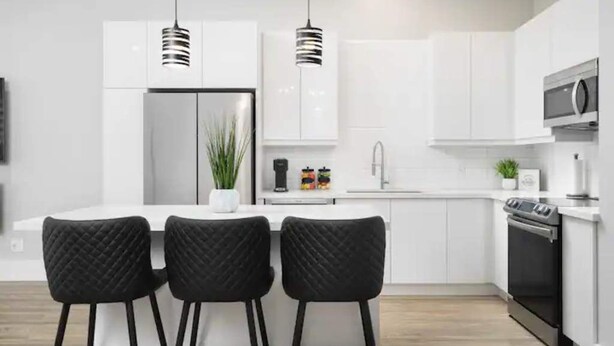 Eine moderne Küche mit weißen Schränken, einer zentralen Kochinsel mit Barhocker, Pendelleuchten, einem Kühlschrank, einer Mikrowelle und einem Backofen.