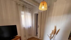 Un salon doté d’un plancher en bois, d’un téléviseur posé sur un meuble en bois, d’un luminaire suspendu et d’une petite table ornée de bougies et de plantes séchées.