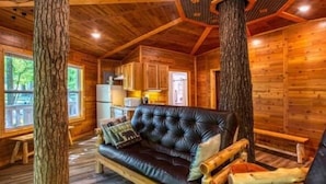 A log cabin interior with a leather sofa, wooden walls, and a large tree trunk centerpiece. - Tree House Deluxe Cabin w/bath - Glamping with real trees growing in the center of the cabin! (Manchester)