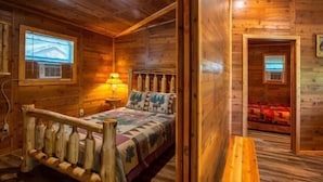 A wooden cabin room with a bed, a nightstand, a lamp, and a window. - Tree House Deluxe Cabin w/bath - Glamping with real trees growing in the center of the cabin! (Manchester)