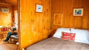A wooden cabin room with a bed, a chair, and a small table. - Deluxe Cabin (w/ Bathroom) - Better than a motel! Enjoy camping in comfort! (Manchester)