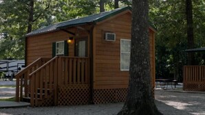 A small wooden cabin with a porch and a staircase, surrounded by trees. - Deluxe Cabin (w/ Bathroom) - Better than a motel! Enjoy camping in comfort! (Manchester)