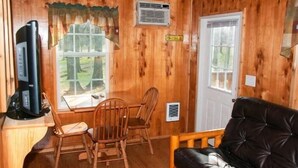 A wooden cabin interior with a dining table, two chairs, a black leather couch, a television, and a window with curtains. - Deluxe Cabin (w/ Bathroom) - Better than a motel! Enjoy camping in comfort! (Manchester)