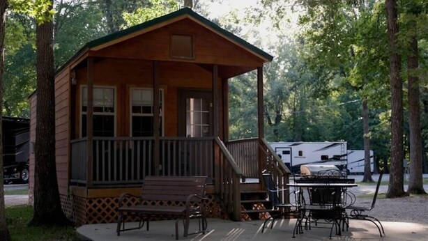 A wooden cabin with a porch, a bench, and a table with chairs. - Deluxe Cabin (w/ Bathroom) - Better than a motel! Enjoy camping in comfort! (Manchester)