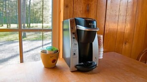 A Keurig coffee maker on a wooden table with a cup of coffee and a pod holder. - Deluxe Cabin (w/ Bathroom) - Better than a motel! Enjoy camping in comfort! (Manchester)