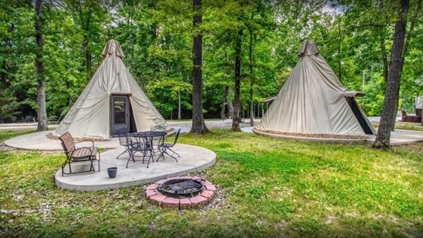 Two tents with attached chairs and a table, a fire pit, and a paved area surrounded by trees.