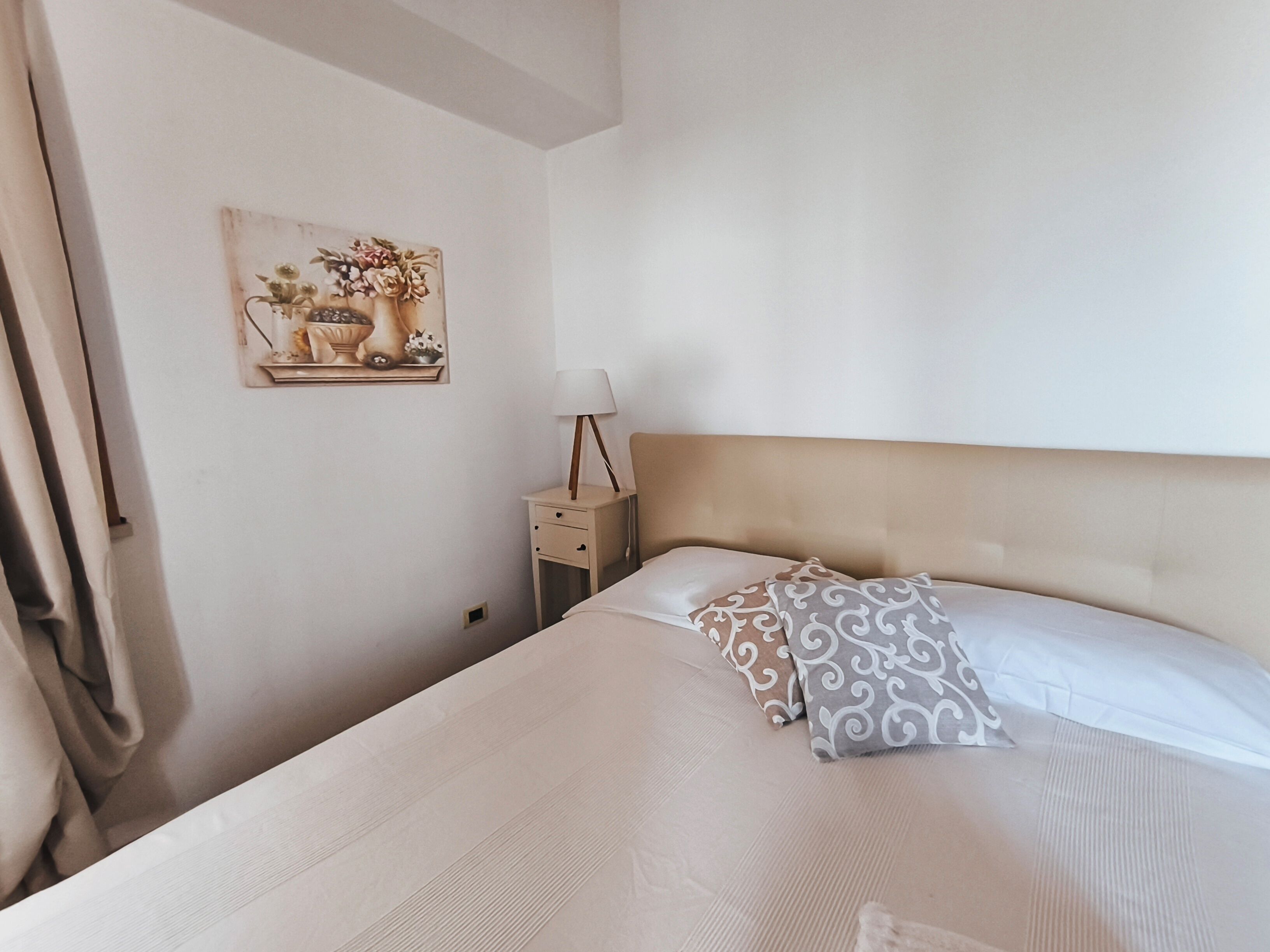 Ein ordentlich bezogenes Bett mit einem gemusterten Kissen, ein Nachttisch mit einer Lampe und ein an der Wand hängendes Kunstwerk.