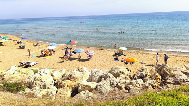 A beach with people sunbathing and swimming, umbrellas, and a rocky foreground. - Avola Opera House (Avola)