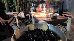 An outdoor dining area with a table set for two under a canopy, featuring a bowl of mussels and bread, wine glasses, and a water bottle. - Avola Opera House (Avola)
