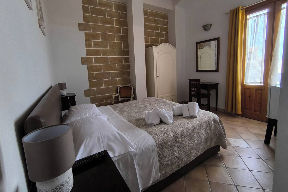 A bedroom with a brick wall, a bed with white linens, a desk, a chair, a mirror, and a window with curtains.