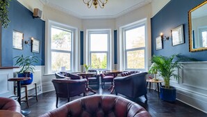 A room with dark wood flooring, leather armchairs, a chandelier, and large windows with a view of trees. - Bourne Hall Country Hotel (Shanklin)
