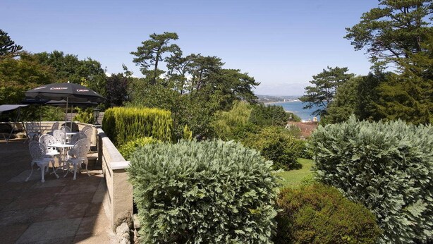 An outdoor seating area with tables and chairs, surrounded by lush greenery and overlooking a coastal view. - Bourne Hall Country Hotel (Shanklin)
