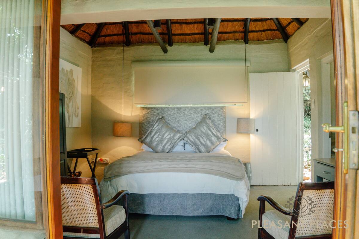 Un dormitorio con cama, dos sillas, una mesita de noche y una puerta que da a otra habitación.