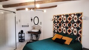 A bedroom with a bed, a desk, a chair, a hanging light fixture, and a shower area. - Les Gîtes de La Roche Élie-chez Théophile (ROM)