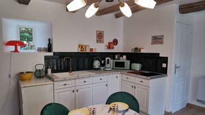 A modern kitchen with white cabinets, a black countertop, and a wooden ceiling. There is a dining table with chairs and a small orange stool. - Les Gîtes de La Roche Élie-chez Théophile (ROM)