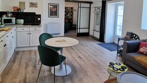 A modern kitchen with white cabinets, a black countertop, and a green chair. There is a dining table with a white tablecloth and a yellow rug. - Les Gîtes de La Roche Élie-chez Théophile (ROM)