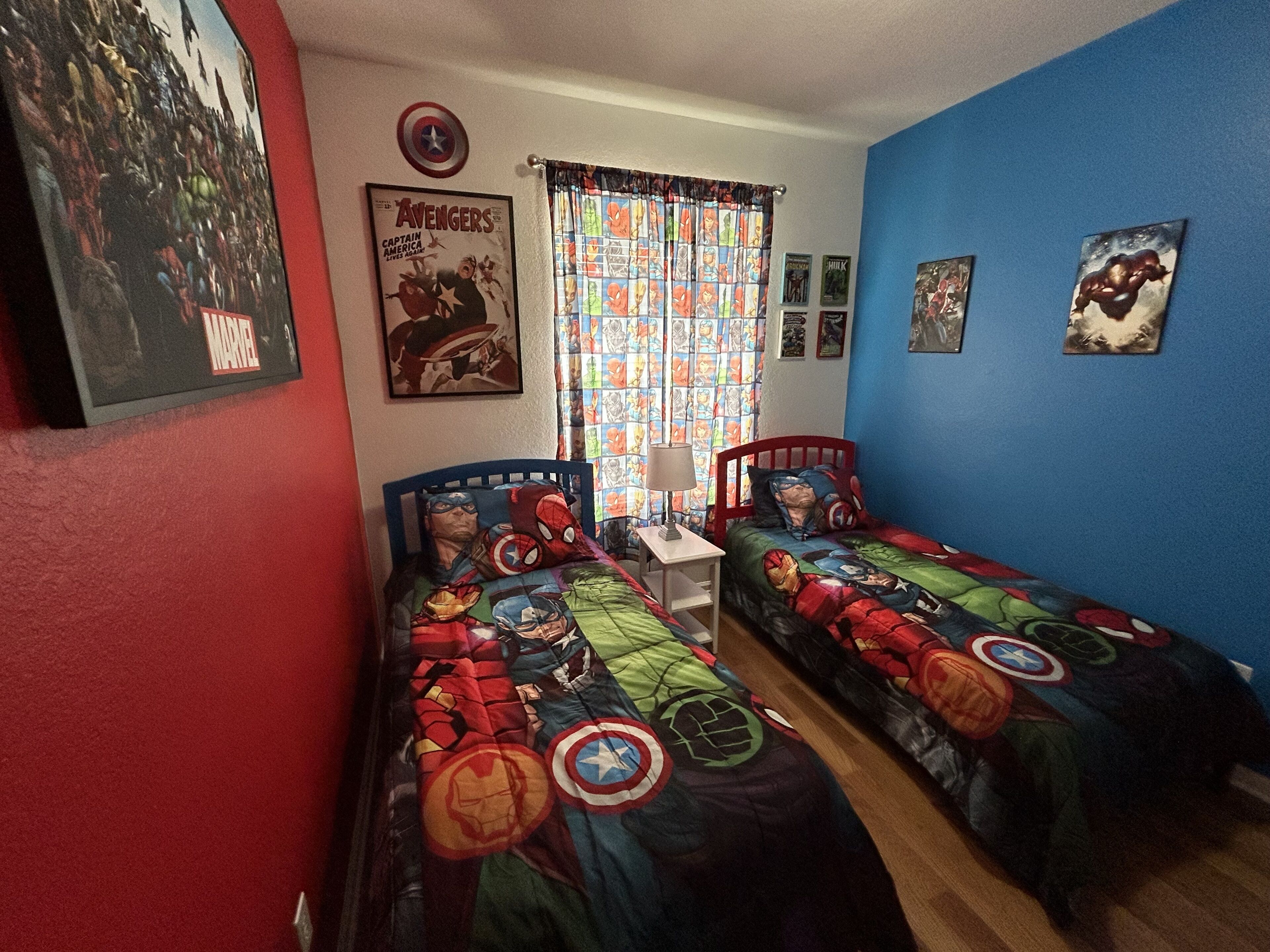 A room with two beds, each covered with superhero-themed bedding. There is a poster of the Avengers on the wall, a window with patterned curtains, and a small table with a lamp.