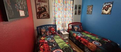 A room with two beds, each covered with superhero-themed bedding. There is a poster of the Avengers on the wall, a window with patterned curtains, and a small table with a lamp.