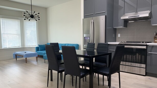 Una cocina moderna con armarios oscuros, electrodomésticos de acero inoxidable y un comedor con sillas negras y una mesa de vidrio.