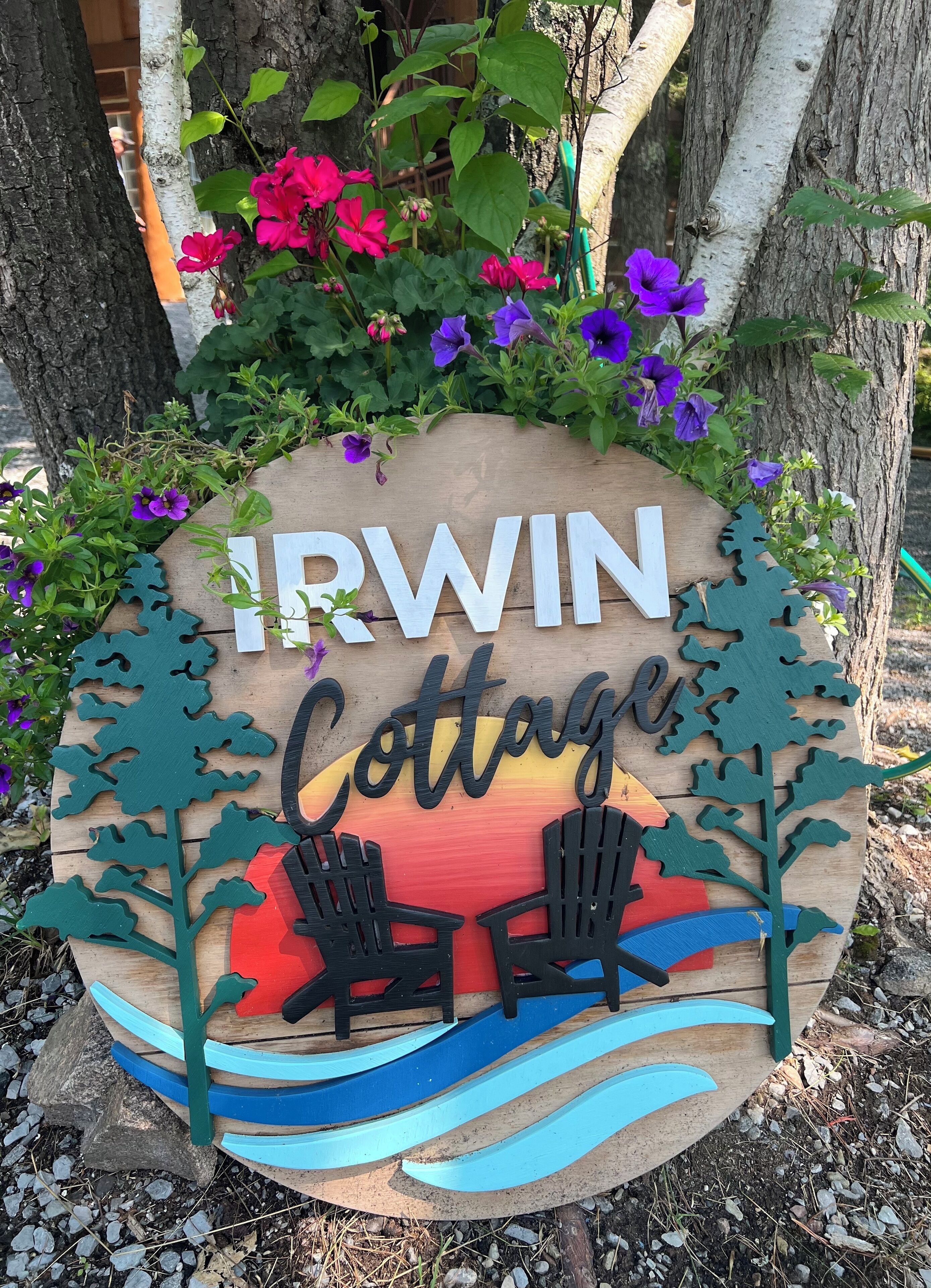 Un panneau en bois indiquant « IRWIN Cottage », ainsi que deux chaises Adirondack, le tout entouré de verdure et de fleurs.