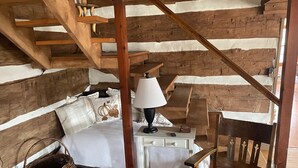 A rustic room with a bed, a desk, a chair, and a staircase. - Oak Haven 1827 Cabin Rental (Union Township)