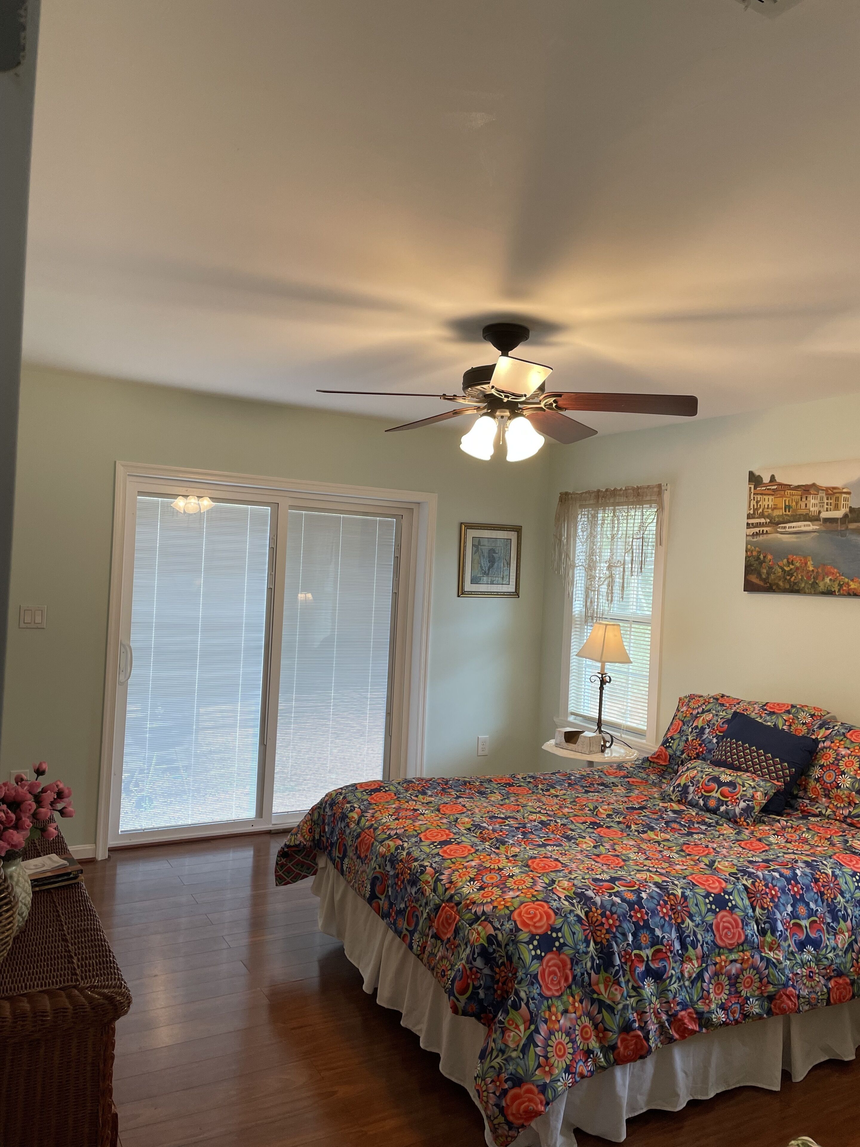 A bedroom with a ceiling fan, a bed with a floral bedspread, a window with blinds, and a painting on the wall.