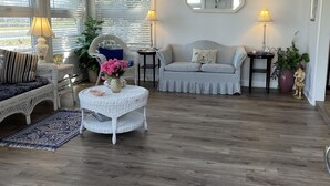 Een ruime woonkamer met een hoge plafond, een plafondventilator en een groot raam met zonwering. Er is een gestreepte bank, een witte rotan stoel en een kleine tafel met een bloevas.