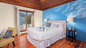 Un dormitorio con techo de madera, una cama con ropa de cama blanca, una pared azul con motivos de hojas, una mesita de noche con una lámpara y una silla con un cojín.