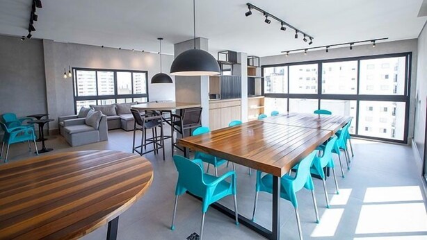 A modern interior with a long wooden table, blue chairs, a sofa, and large windows offering views of buildings. - Studio Sao Paulo PUC Allianz Park 901WB (São Paulo)