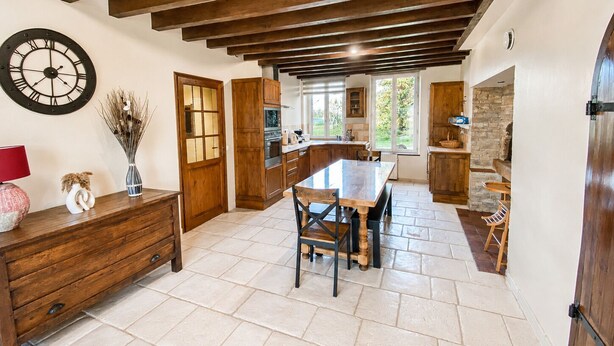 Une cuisine avec des poutres en bois, une table à manger, des chaises, une horloge et un vase contenant des plantes séchées.