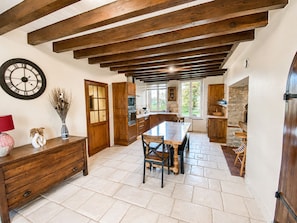 A kitchen with wooden beams, a dining table, chairs, a clock, and a vase with dried plants.