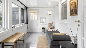 A modern, minimalist living room with a grey sofa, a wooden bench, and a small table. There are windows on both sides, a door, and a framed picture on the wall.
