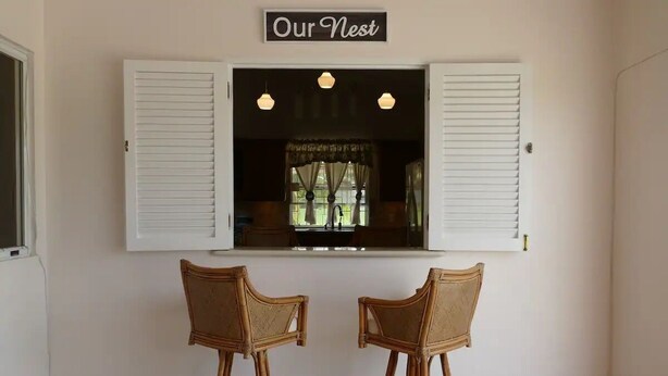 Ein Zimmer mit Fenster, ausgestattet mit weißen Fensterläden, zwei Korbstühlen und einem Schild über dem Fenster mit der Aufschrift 'Our Nest'.