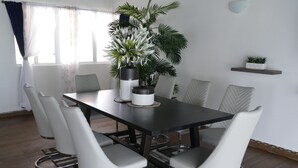 Ein Essbereich mit einem schwarzen Tisch, sechs weißen Stühlen, einem Blumentopf und einem kleinen Regal mit einer Pflanze.