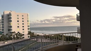 A balcony with a view of the ocean, a tennis court, and a building in the background.
