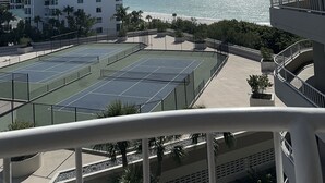 A view of a tennis court with a pool, a beach, and a high-rise building in the background.