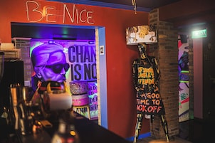 Una zona de bar con un letrero de neón que dice "BE NICE" y un maniquí vestido con un atuendo colorido.
