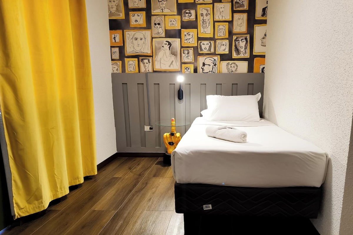 Un petit lit bien fait, avec un oreiller blanc et une couverture noire matelassée, un rideau jaune et un mur décoré de portraits en noir et blanc encadrés.