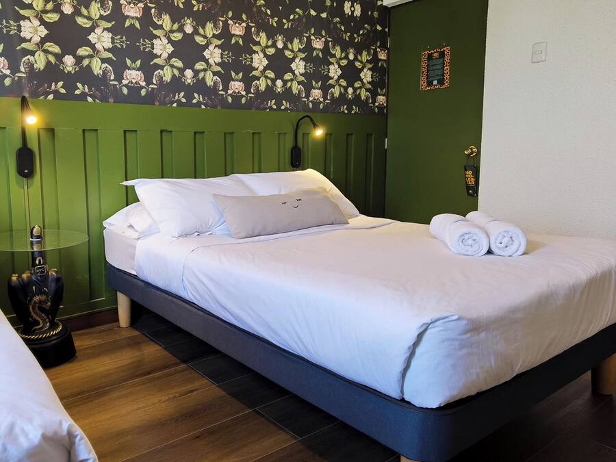 Ein ordentlich bezogenes Bett mit weißen Leinen und zwei aufgerollten Handtüchern, eine grüne Wand mit Blumenwallpaper und eine Wandlampe.