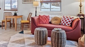 Una sala de estar acogedora con un sofá rojo, dos otomanas de mimbre, una mesa de comedor de madera con sillas amarillas y vistas a un paisaje nevado a través de las ventanas.