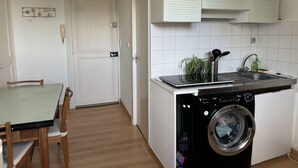 A compact kitchen with a washing machine, a dining table with chairs, and a white door. - T1 bis Proche Viarme - Longue Durée (Nantes)