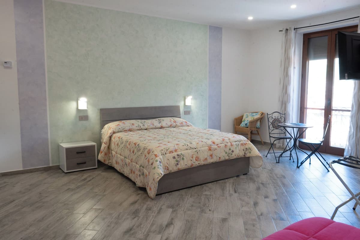 Un dormitorio con cama, mesitas de noche, una silla, una pequeña mesa y una ventana con cortinas.