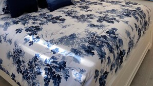 A bedroom with a large bed, a nightstand with a lamp, a window with blinds, and a painting of the sea on the wall.